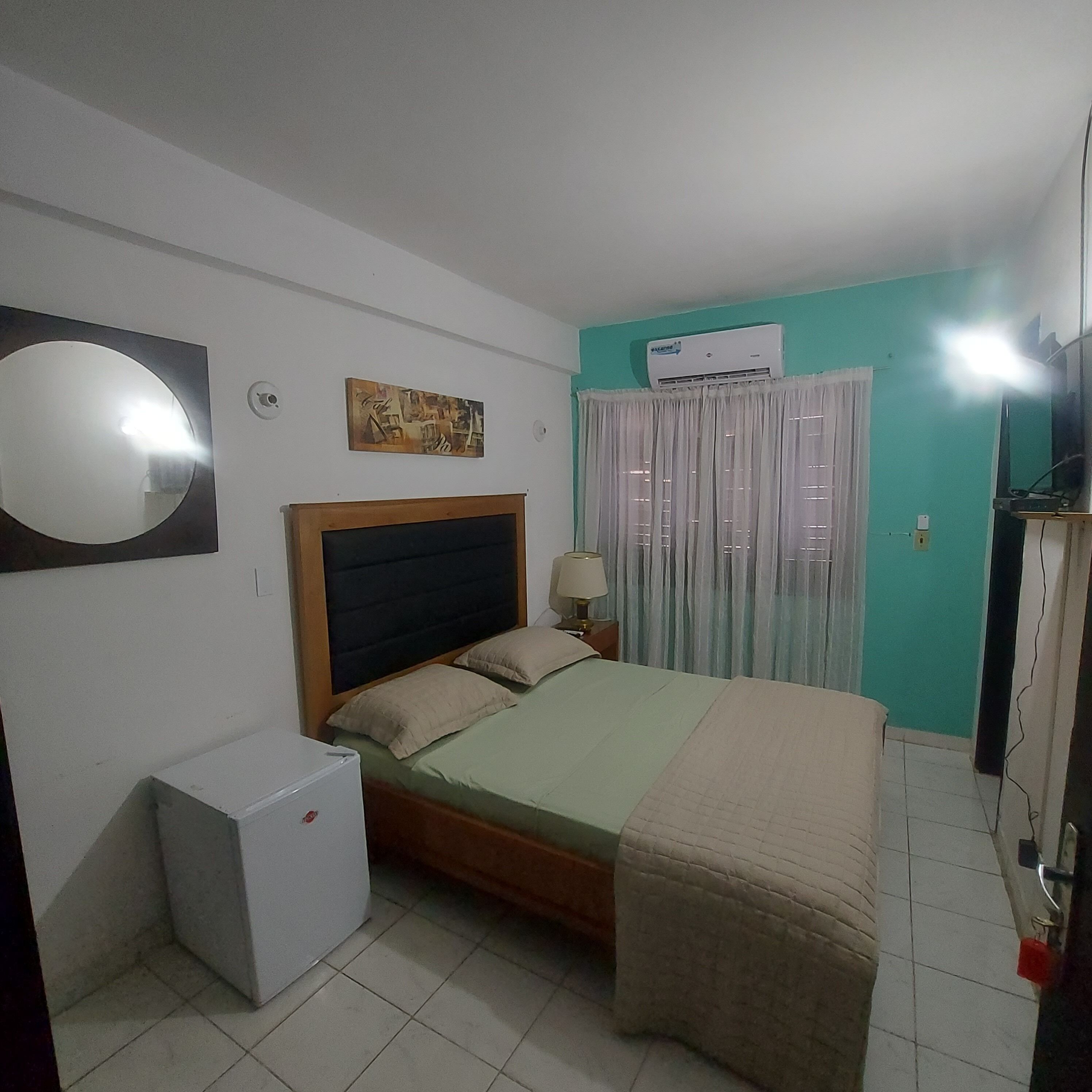 Un dormitorio con cama, un refrigerador pequeño, aire acondicionado instalado en la pared, televisor y ventana con cortinas.