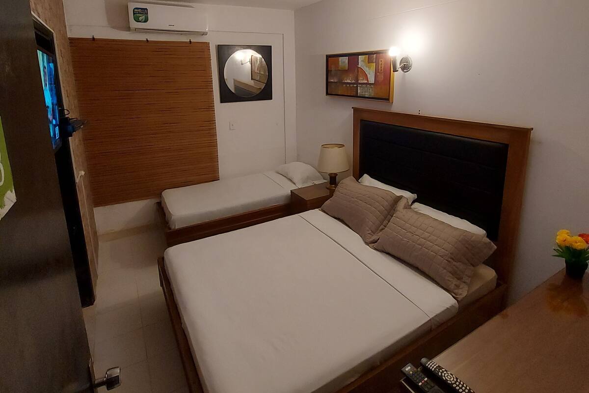 A hotel room with two beds, a wooden headboard, a wall-mounted air conditioner, a mirror, a framed artwork, and a bedside lamp.