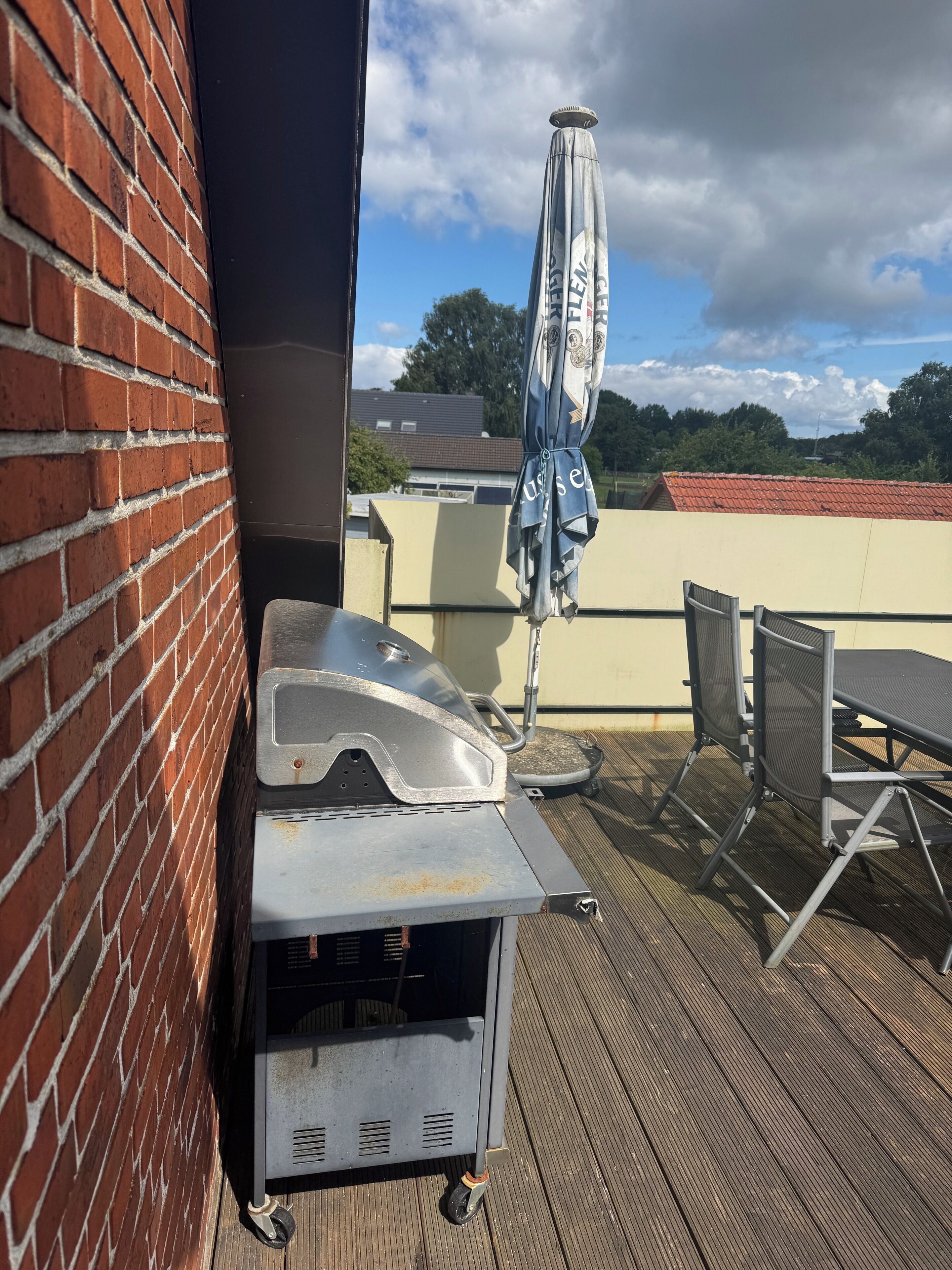 Un barbecue, une table d’extérieur, des chaises et un parasol sur une terrasse en bois.