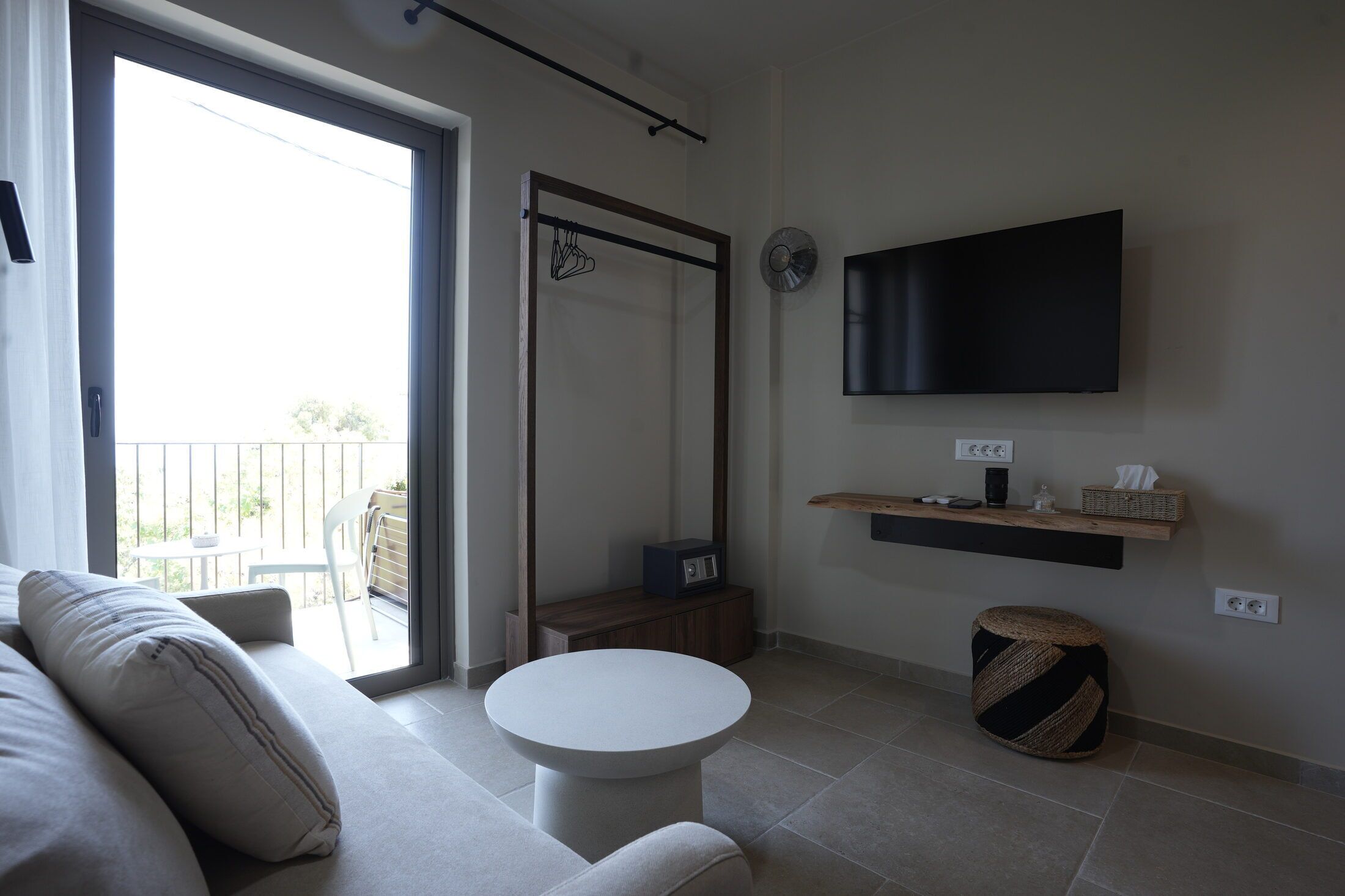 A modern living room with a sofa, a round coffee table, a television mounted on the wall, a small wooden cabinet, and a sliding glass door leading to an outdoor area.