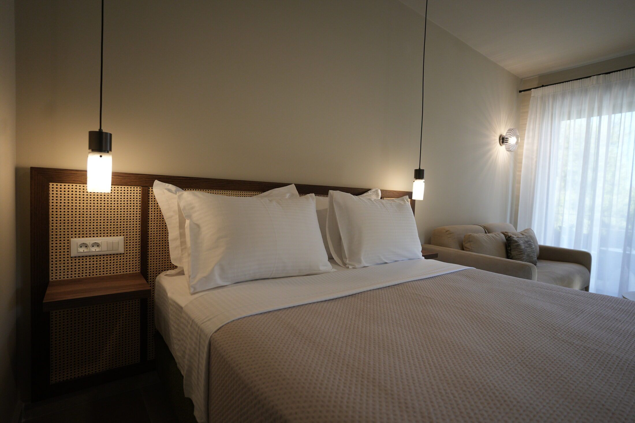 A hotel room with a large bed, bedside tables, a wall socket, a sofa, and a window with curtains.