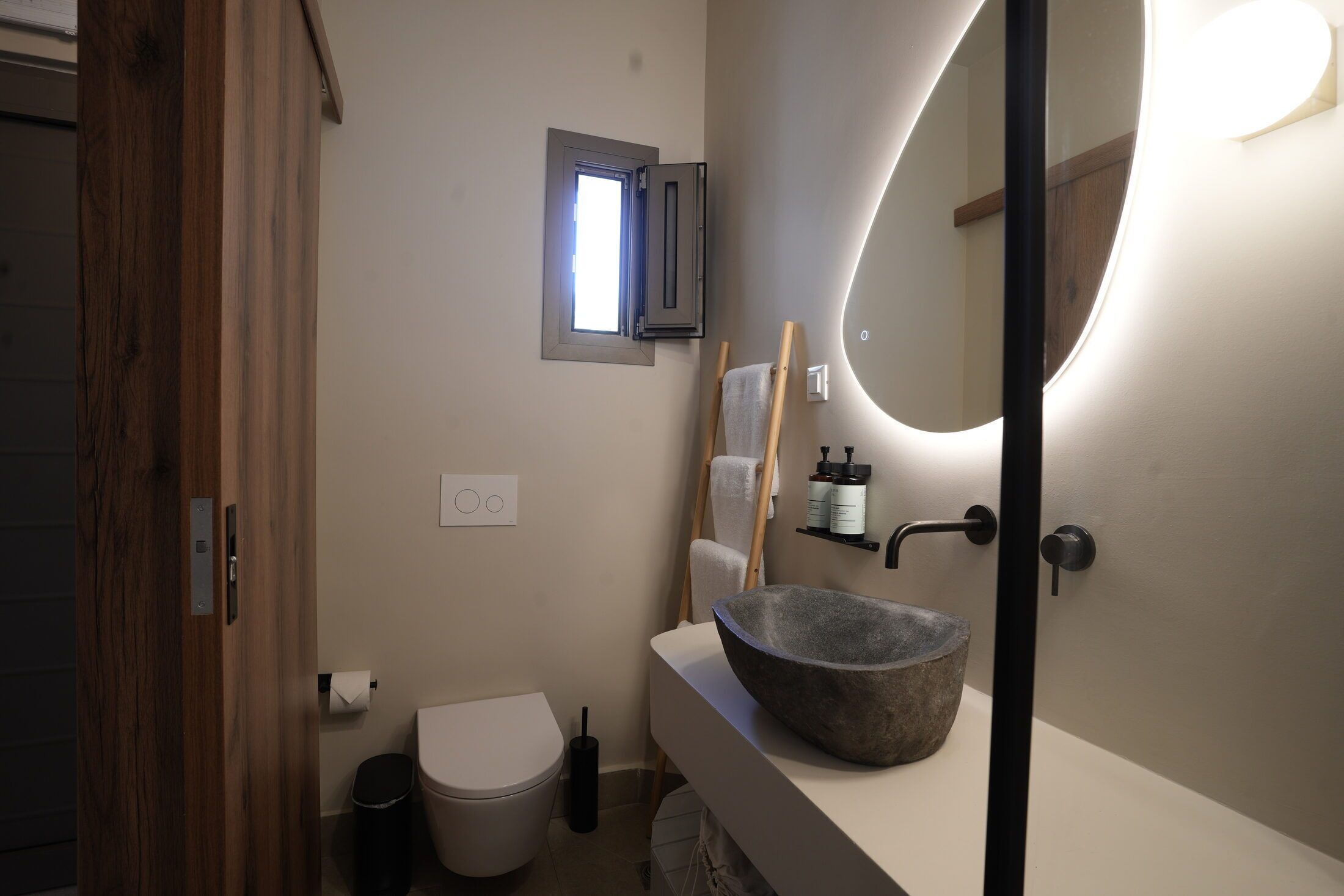 Un baño con puerta de madera, inodoro blanco, lavabo de piedra, pequeña ventana y espejo con luz.