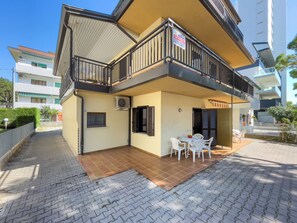 A two-story building with a balcony, a tiled patio, and a sign indicating a rental property. - Lignano Flat Steps From the Beach (Lignano Sabbiadoro)