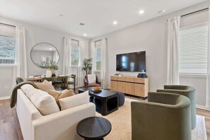 A modern living room with a sofa, coffee table, armchairs, a TV, and a dining area. - Luxe Family-Friendly 3BR with Game Room & Fire Pit (Houston)