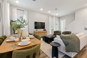 A modern living room with a dining area, a flat-screen TV, and a staircase. - Luxe Family-Friendly 3BR with Game Room & Fire Pit (Houston)