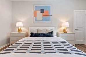 A bedroom with a bed, two bedside tables, a nightstand, a door, and a wall-mounted abstract painting. - Luxe Family-Friendly 3BR with Game Room & Fire Pit (Houston)