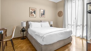Ein Schlafzimmer mit einem Bett, zwei Nachttischlampen, einem Sessel, einem Schreibtisch und einem Fenster mit Vorhängen.