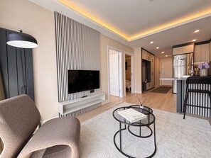 A modern living room with a flat-screen TV, a sofa, a coffee table, and a kitchen area with a refrigerator and cabinets. - Chic & Cozy 2BR – Nişantaşı (Istanbul)
