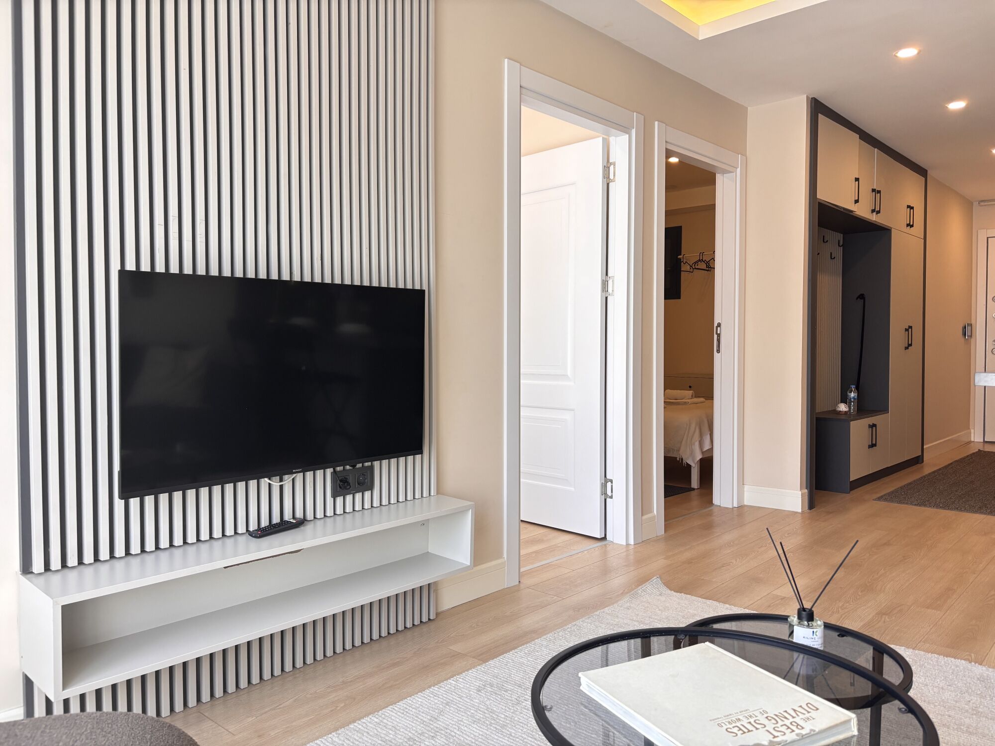 Un salon moderne avec un téléviseur à écran plat fixé au mur, une table basse en verre et une porte donnant sur une chambre à coucher.