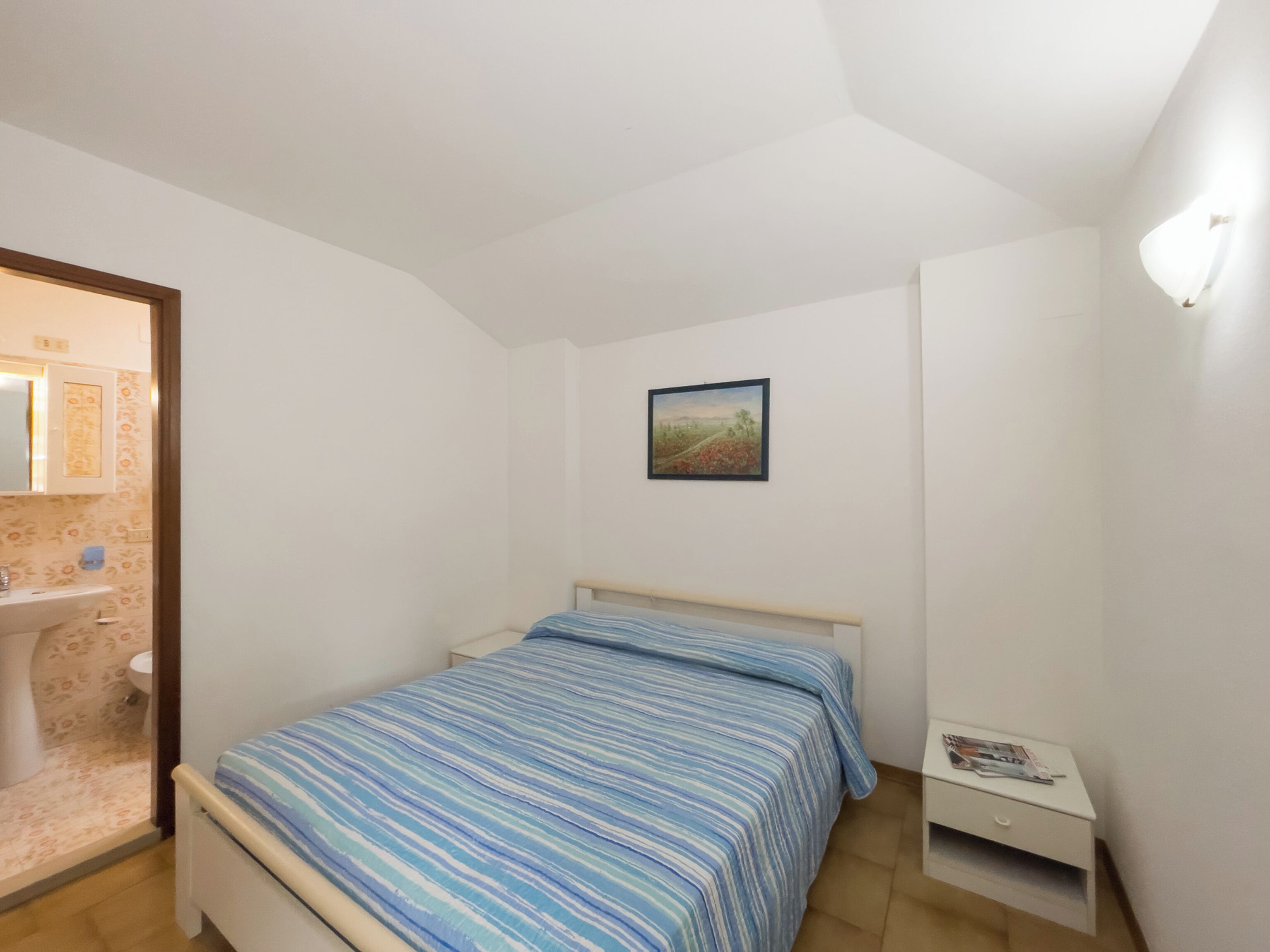 A bedroom with a bed, a nightstand, a painting, and a bathroom visible through an open door.