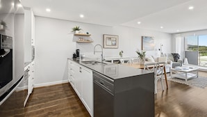 Una cocina moderna con armarios blancos, lavavajillas integrado y una isla grande. Hay una zona de comedor con una mesa y sillas blancas, y una sala de estar con un sofá y una mesa de centro.