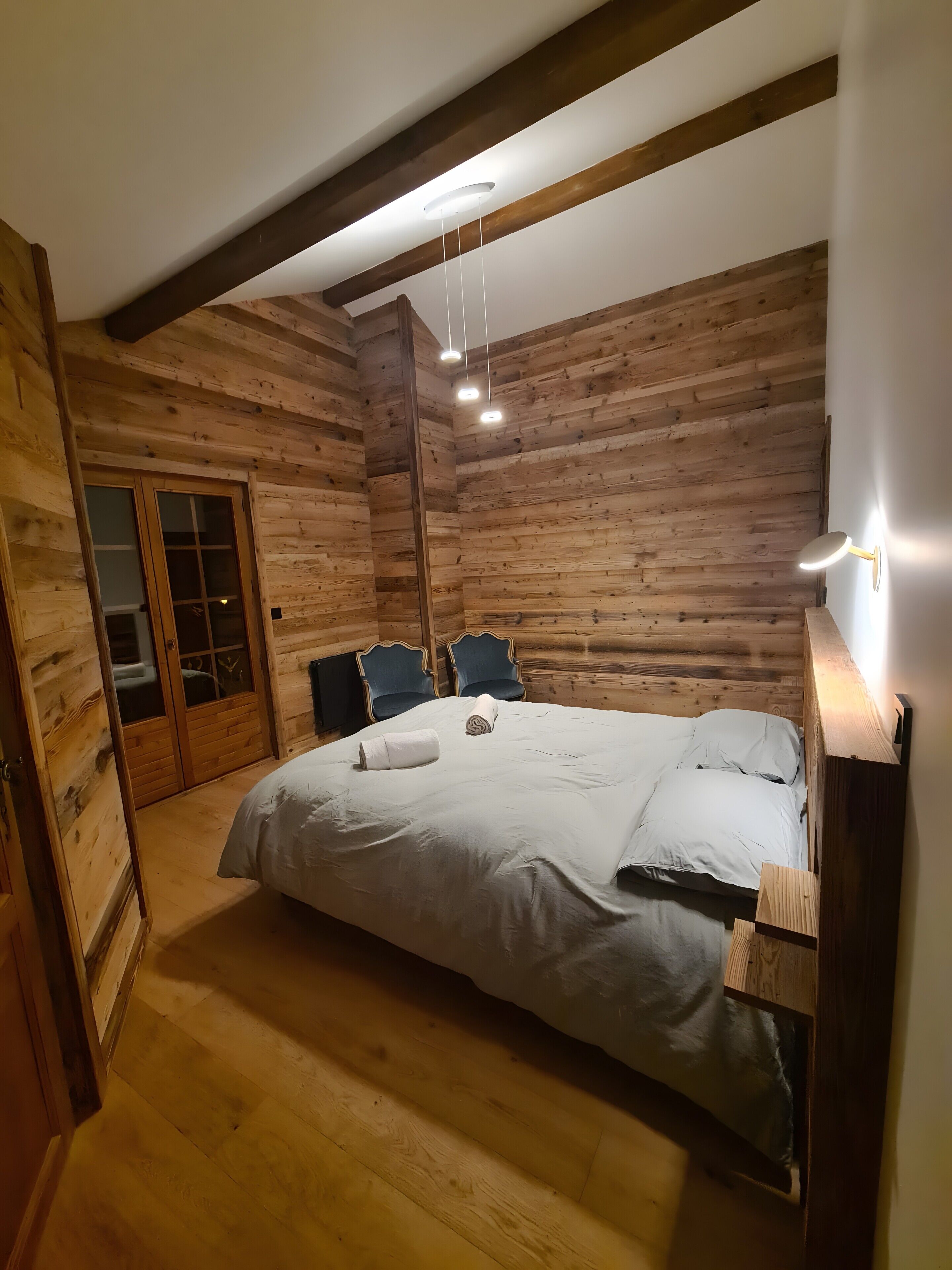 Een slaapkamer in een blokhut met een bed, twee stoelen, een houten plafond en een deur.