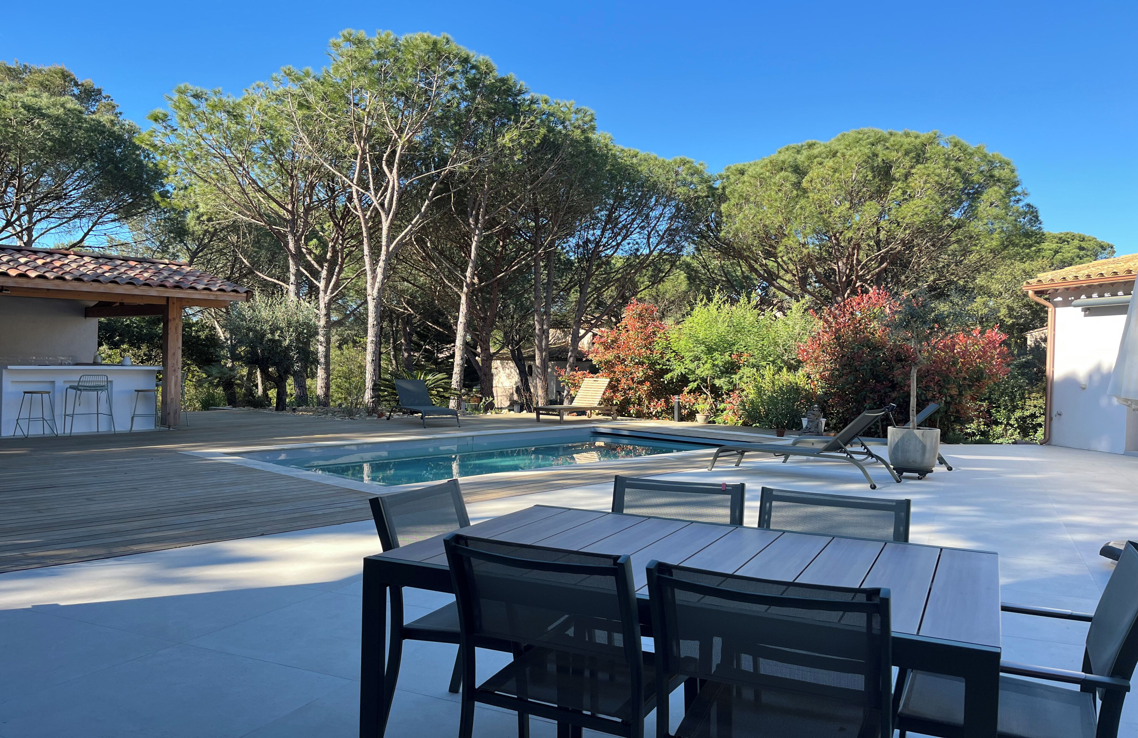A poolside area with a wooden deck, outdoor seating, and a clear view of trees and a building in the background.