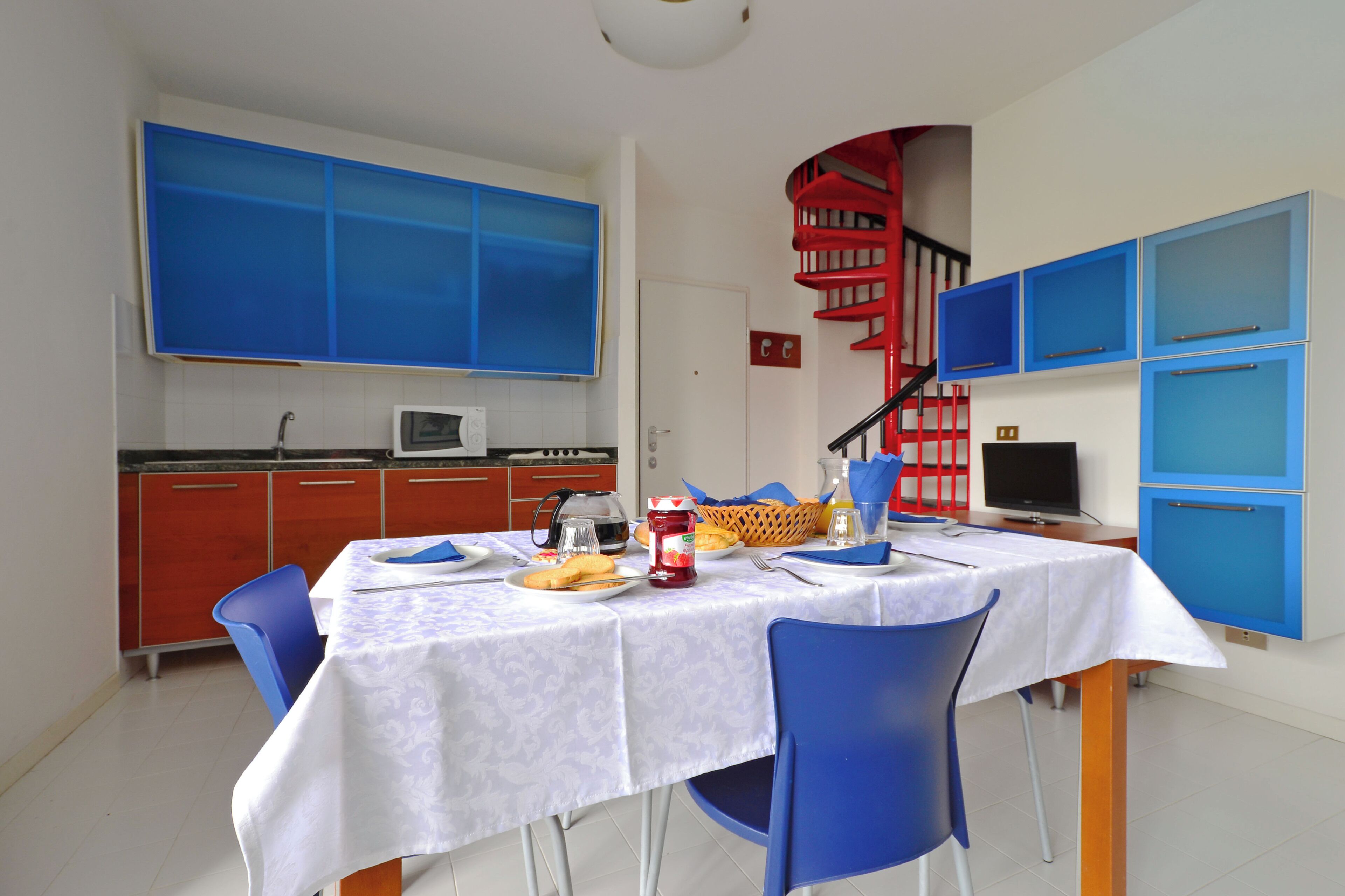 A dining area with a table set for two, a kitchen in the background, and a red spiral staircase.