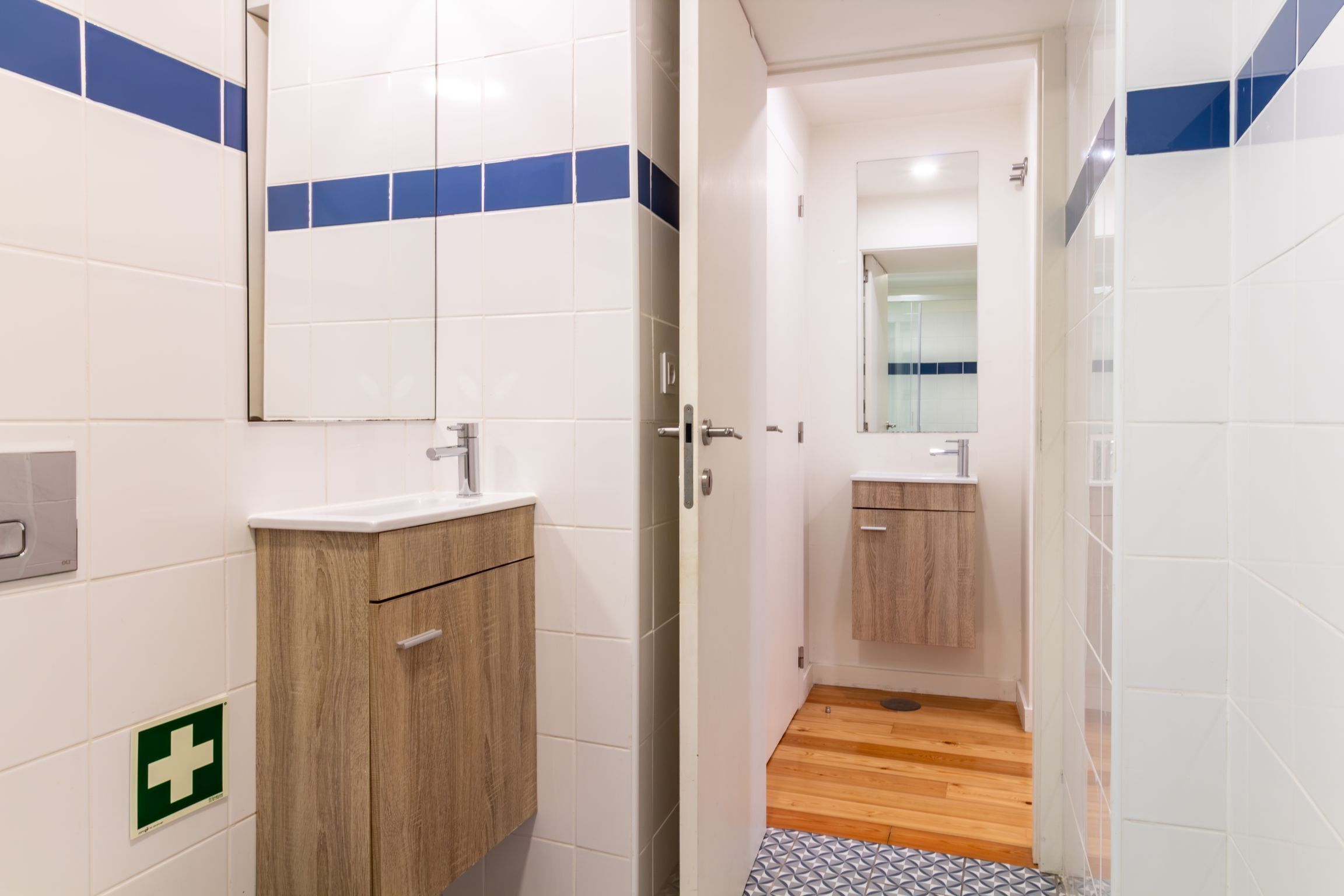 A bathroom with white tiles, a wooden vanity, a mirror, and a green emergency exit sign.