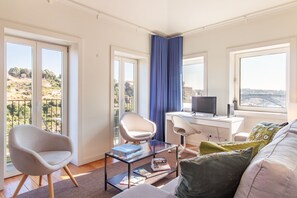 A modern living room with a glass coffee table, a white sofa, a desk with a computer, and a view of a cityscape through large windows. - Corticeira Living (Porto)