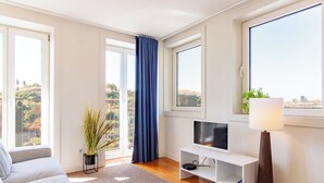 A modern living room with a sofa, a TV on a stand, a lamp, and large windows with curtains. - Corticeira Living (Porto)