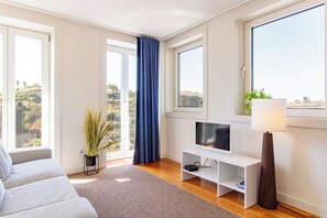 A modern living room with a sofa, a TV on a stand, a lamp, and large windows with curtains. - Corticeira Living (Porto)