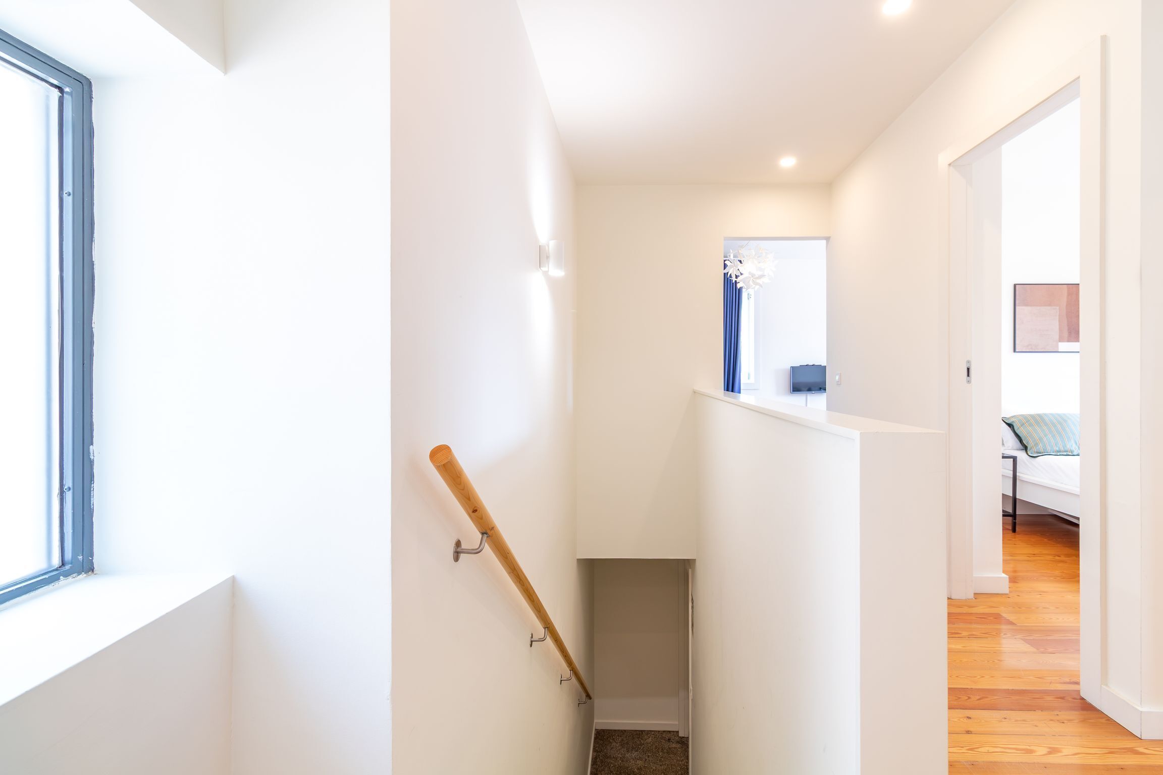 A narrow hallway with a wooden handrail, a small enclosed space, and a bedroom with a bed and a blue chair.