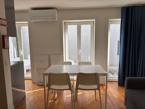 A room with a white table, two chairs, a window with curtains, an air conditioner, and a door. - Corticeira Living (Porto)