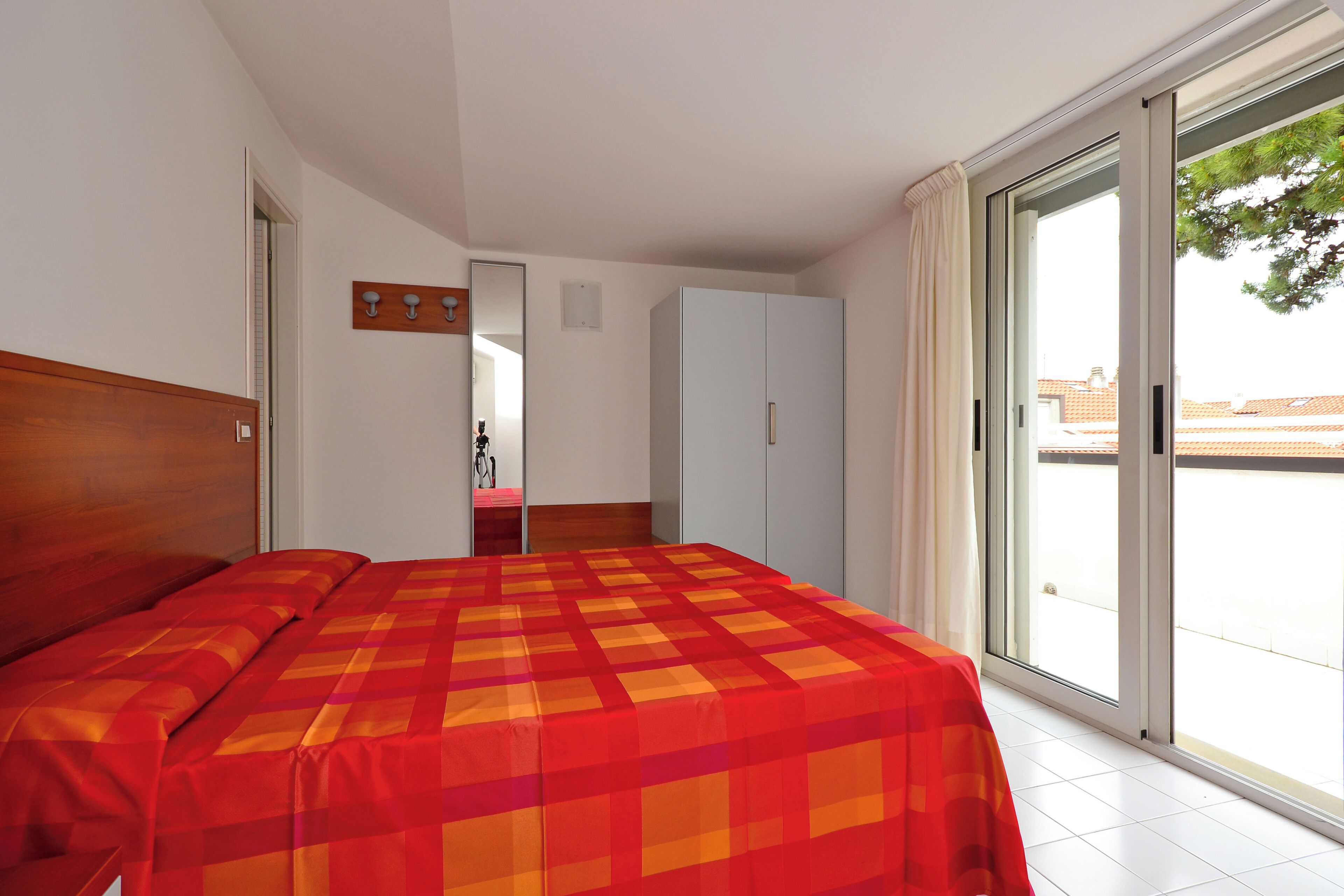 Quarto com cama, guarda-roupa, espelho e porta de vidro deslizante para uma varanda.