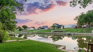 A serene lakeside property with a reflective pond, a wooden dock, and a house with a covered porch. - Lakefront!  Fire Pit, Kayaks, Foosball, Sparkling Clean Hot Tub (Granbury)