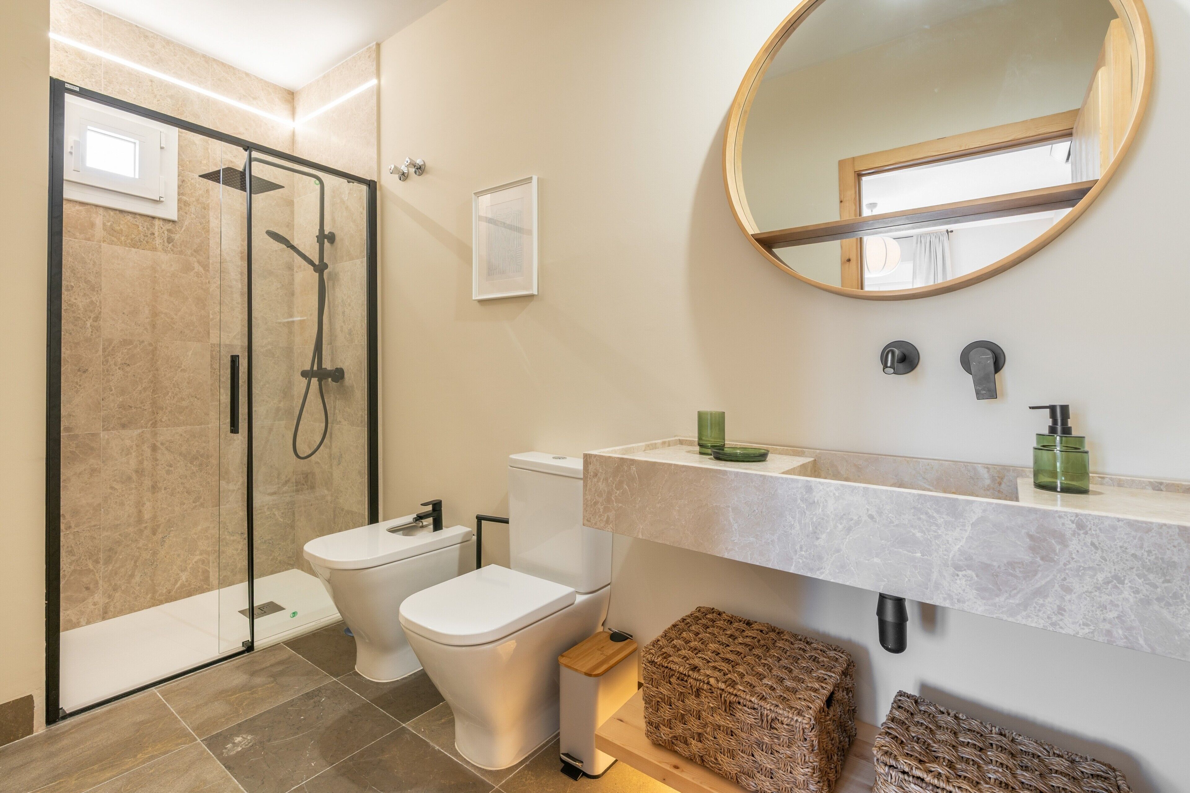 Een moderne badkamer met een glazen douchecabine, toilet, wastafel met een marmeren blad en een ronde spiegel.