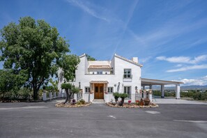 A white building with a wooden door, surrounded by potted plants and a paved driveway. - Casa Rural Huerta Real (Baza)