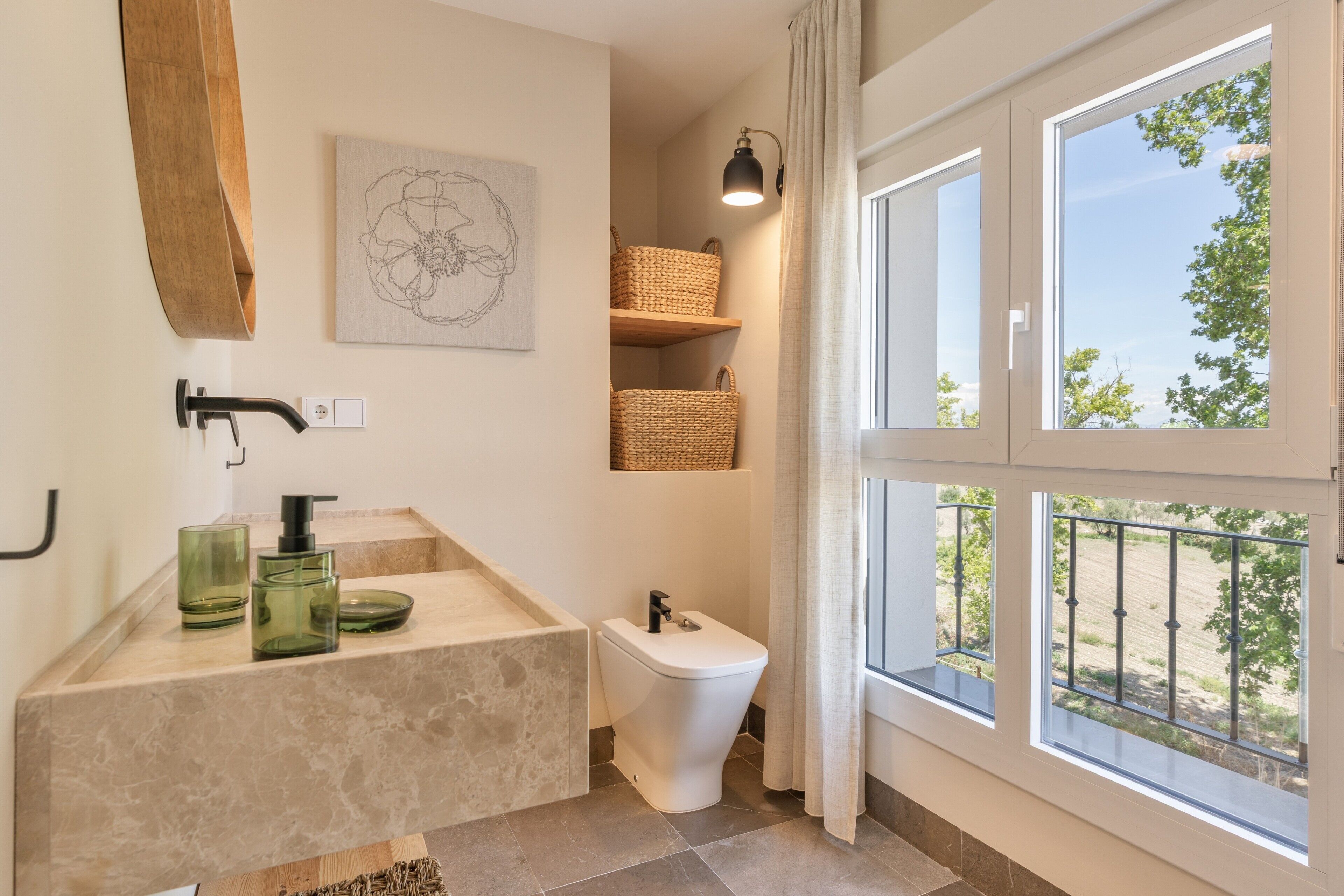 Un baño con lavamanos de mármol, inodoro, cestas de mimbre y ventana con vistas a la vegetación.