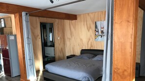 A bedroom with a bed, wooden walls, a mirror, a small desk, and a refrigerator.