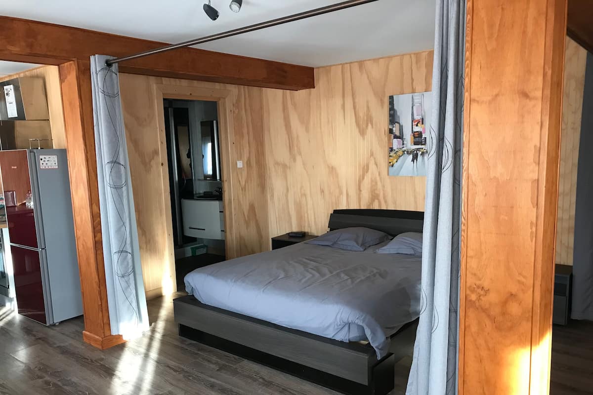 A bedroom with a bed, wooden walls, a mirror, a small desk, and a refrigerator.