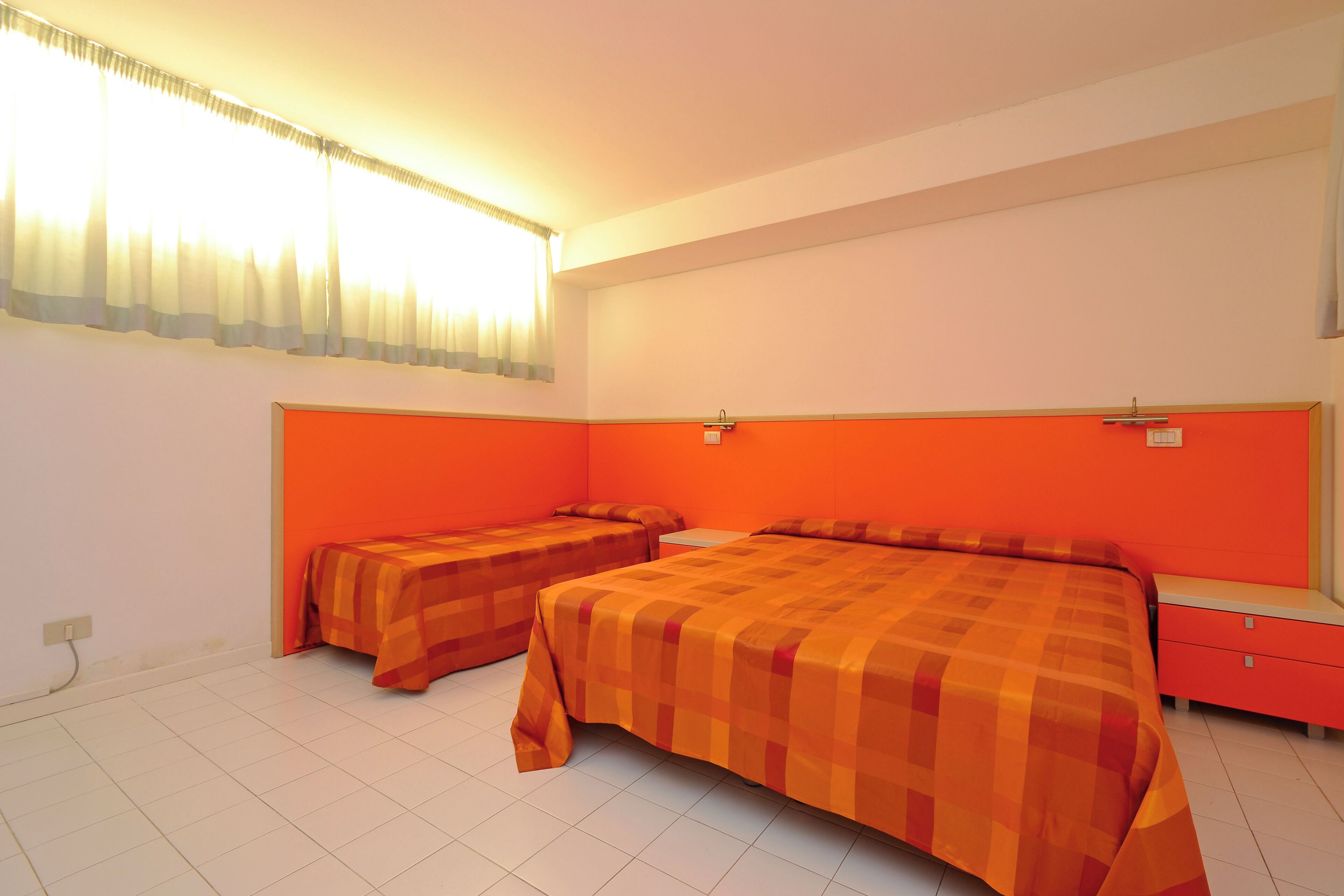 Een kamer met twee bedden, een nachtkastje, een raam met gordijnen en een aan de muur bevestigd lampje.