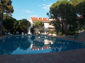 A swimming pool with lounge chairs, trees, and a building in the background. - Sweet Holiday Apartment With Pool (Lignano Sabbiadoro)