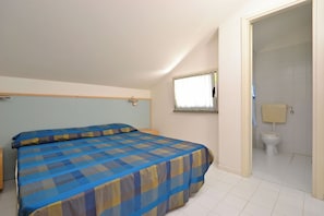 A bedroom with a bed, a nightstand, a window with curtains, and a bathroom with a toilet and sink. - Sweet Holiday Apartment With Pool (Lignano Sabbiadoro)