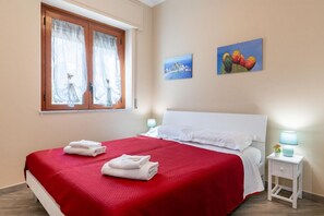 A neatly made bed with a red bedspread, white pillows, and two folded white towels. There is a window with sheer curtains, a small bedside table with a lamp, and two wall-mounted paintings. - Cozy and bright apartment of approximately 40 square meters, perfect for four people, located on the ground floor of a 1980s building.The property enjoys an excellent location in Giardini Naxos, just 5 minutes from the beach. Nearby amenities such as bar (Naxos)