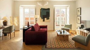 A living room with a sofa, coffee table, and chairs, a desk with a lamp, and a large window with a view of buildings. - Ando Living - Fanqueiros House (Lisbon)