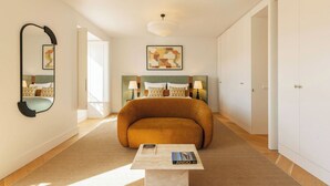 A hotel room with a curved sofa, a bed with patterned bedding, a bedside table with a lamp, a large mirror, and abstract artwork on the wall. - Ando Living - Fanqueiros House (Lisbon)