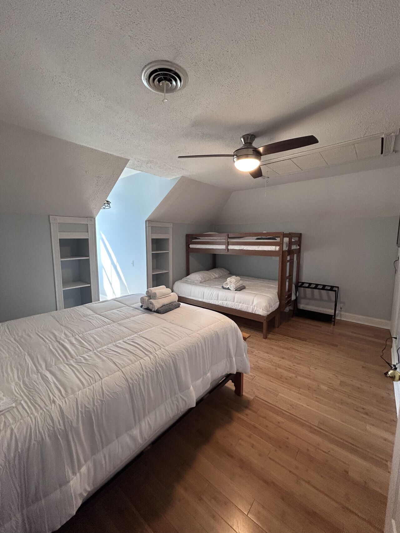 Une chambre avec un lit superposé, un lit simple, un ventilateur de plafond et une lucarne.