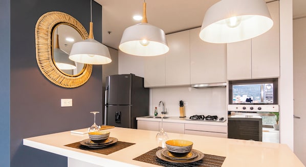 A modern kitchen with a central island, pendant lights, a refrigerator, stove, and a large mirror. - Apartamento Urban Studios (Medellín)