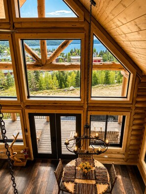 A log cabin interior with large windows, a wooden dining table with a vase of flowers, and a chandelier.