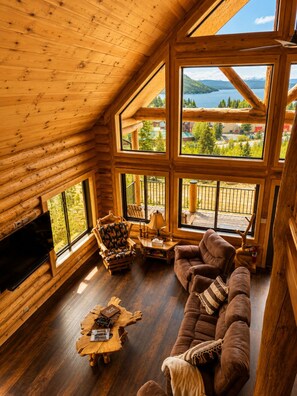 LâintĂ©rieur dâune cabane en rondins, avec une grande fenĂȘtre, un tĂ©lĂ©viseur, un canapĂ© marron, une table basse en bois et un fauteuil ornĂ© dâun motif de renne.