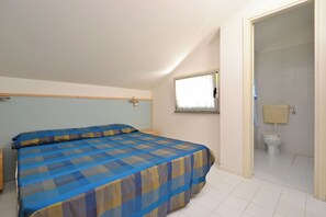 A bedroom with a bed, a nightstand, a window with curtains, and a bathroom with a toilet and sink. - Quiet Living for Your Vacation (Lignano Sabbiadoro)