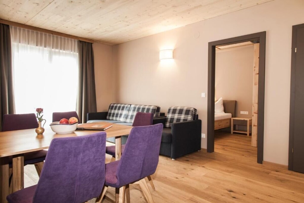 A room with a dining table, purple chairs, a sofa with plaid cushions, a bed, and a small bedside table.