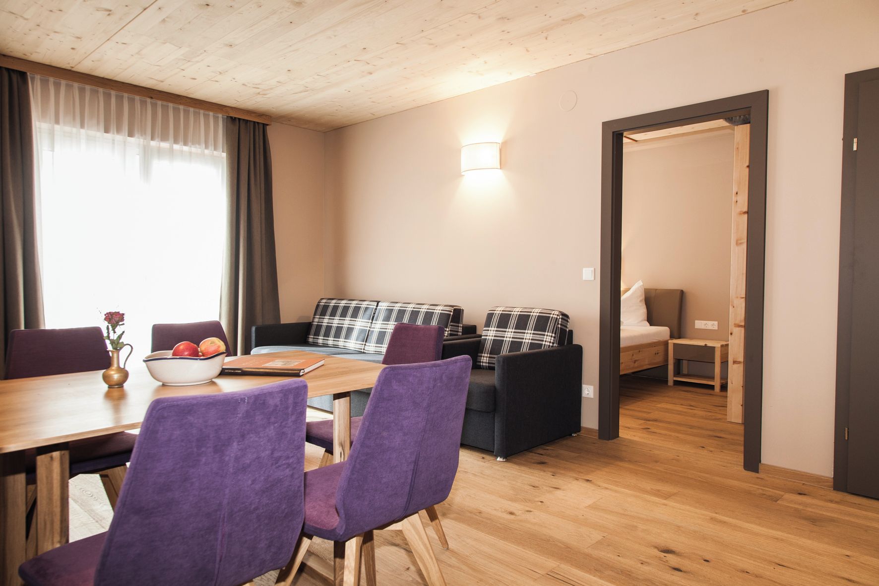 Un salon moderne avec un plafond en bois, une table à manger entourée de chaises violettes, un canapé agrémenté de coussins à carreaux et un lit dans la pièce adjacente.