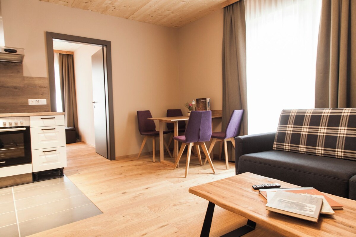 A compact living space with a kitchenette, a dining area with chairs, a sofa, and a table with a remote control and books.
