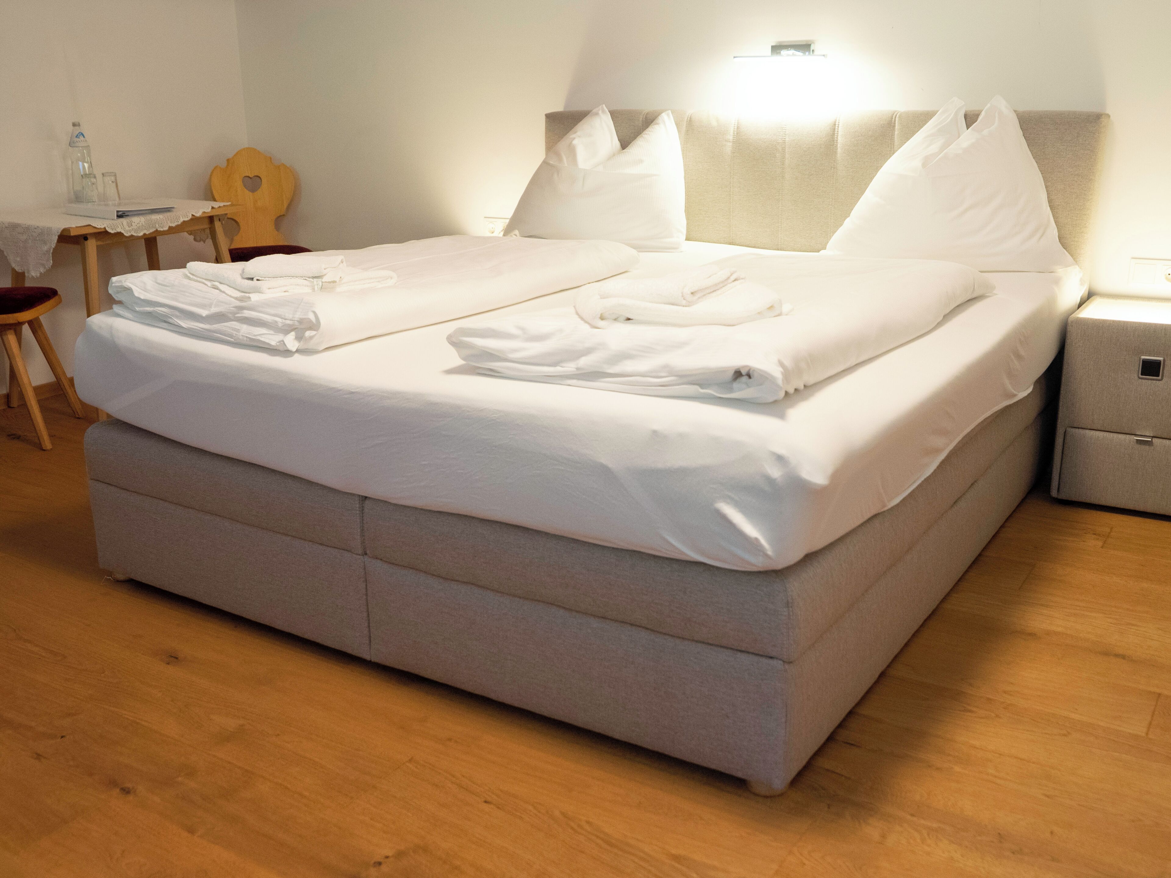 A neatly made bed with pillows and folded towels, a bedside table with a lamp, and a wooden chair.