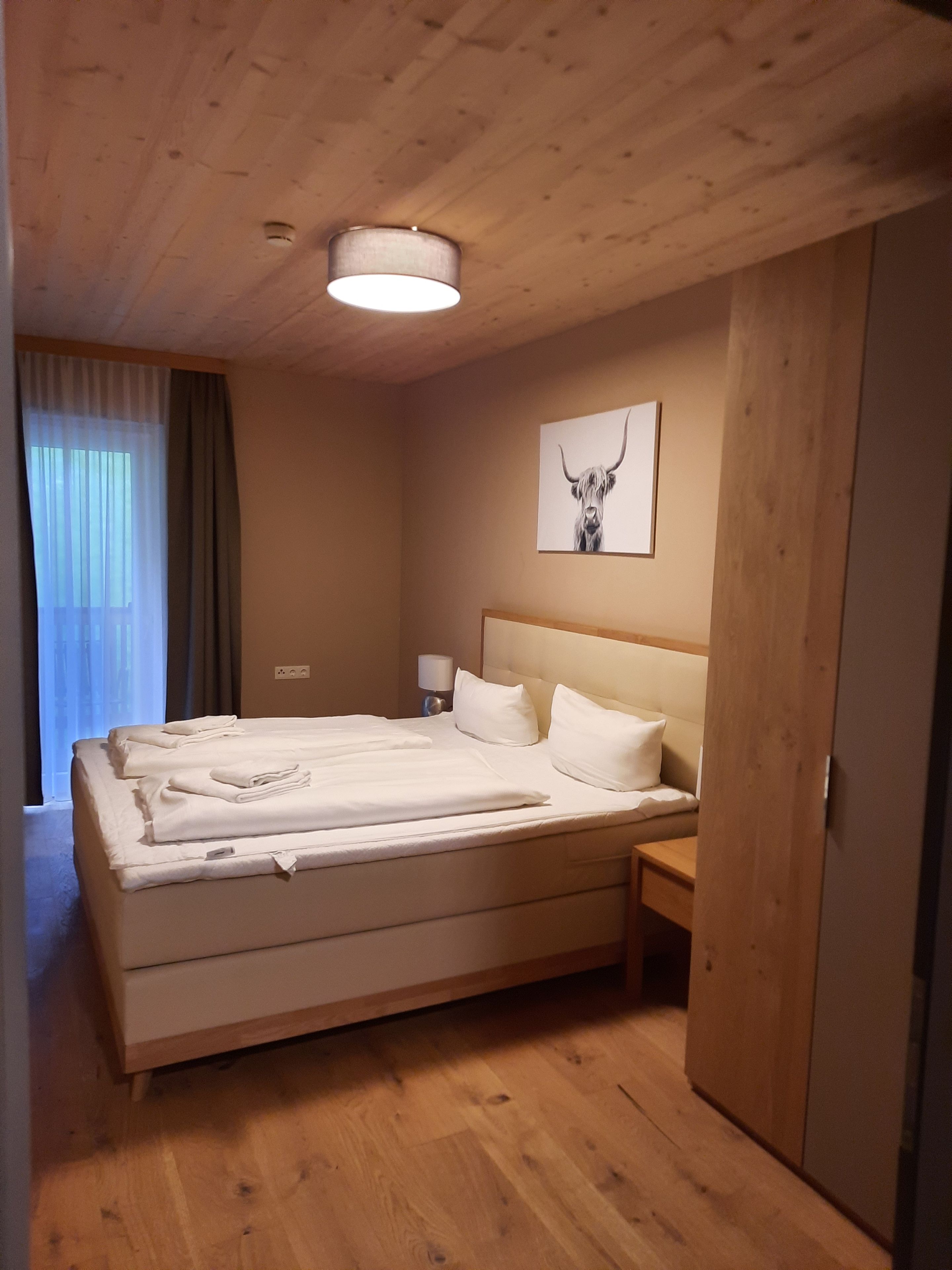 Habitación de hotel con techo de madera, una cama con sábanas blancas, una mesita de noche, una lámpara, un cuadro en la pared con la imagen de una cabeza de toro y una ventana con cortinas.