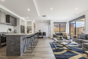 Eine moderne Küche mit einer zentralen Kochinsel, Barhocker, einem Essbereich mit Stühlen, einer Couch und einem großen Fenster mit Blick auf einen Garten.