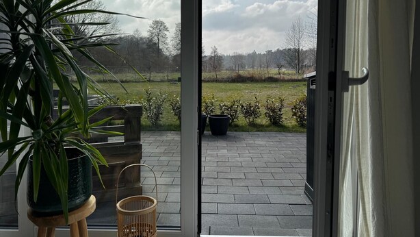 Ein Zimmer mit einer Schiebetür zu einer Terrasse im Freien, mit Blick auf ein Wiesenfeld und Bäume.