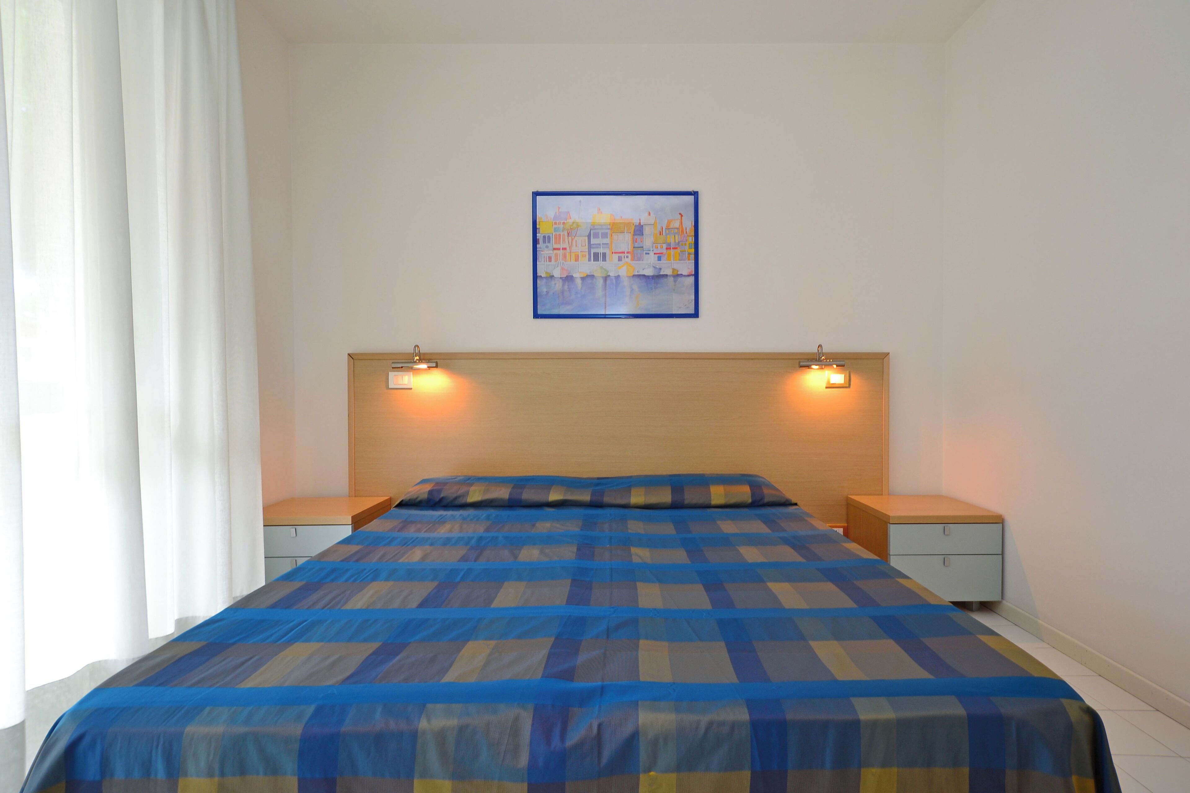 A bedroom with a checkered bedspread, two bedside lamps, a nightstand, a wooden headboard, and a framed picture on the wall.